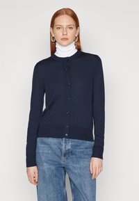 Mornarsko modra jopica iz mehkega materiala, z okroglim izrezom, gumbnim zapiranjem in rebrastim robom. Nosi se čez belo turtleneck.