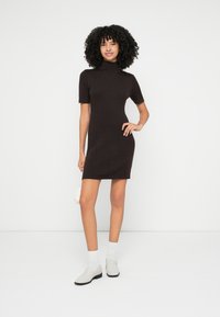 Pletené šaty v tmavohnedej farbe s vysokým golierom a krátkymi rukávmi. Hladká textúra, priliehavý strih, kombinované s bielymi ponožkami a svetlými topánkami.