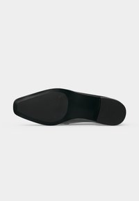 Semelle de chaussure habillée noire avec un bout pointu et un dessous lisse et plat, vue par en dessous sur un fond clair uni.