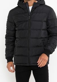 Sort puffy jakke med hætte, der har quiltede sektioner, en frontlynlås og ribbet manchetter. Glat stof med et slankt design.