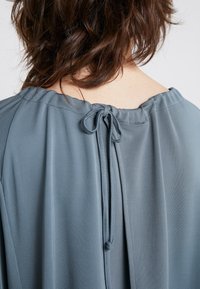Szara bluzka z okrągłym dekoltem, z wiązaniem z tyłu, o miękkiej teksturze materiału i luźnym kroju. Prosty, elegancki design.