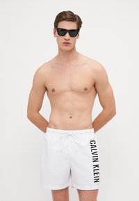 Biele plavky s viazaním v páse, na boku s čiernym logom "CALVIN KLEIN". Model s okuliarmi na slnko, bez trička.