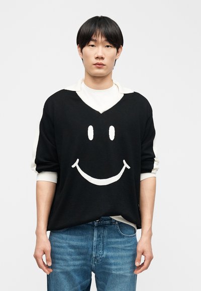 Νεαρός άνδρας με ίσια μαύρα μαλλιά, ντυμένος με μαύρο πουλόβερ που έχει μεγάλο λευκό χαμογελαστό πρόσωπο, λευκό πουκάμισο από κάτω και μπλε τζιν.