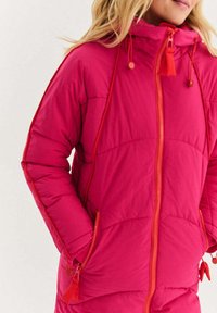 Růžová nadýchaná bunda s celopropínacím zipem, kapucí s nastavitelnými šňůrkami a kontrastními oranžovými akcenty na zipu a švech.