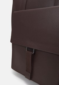 Poche de sac à dos en cuir marron foncé avec fermeture à rabat sécurisée par une sangle en tissu et une boucle métallique, montrant une couture détaillée sur une surface lisse.