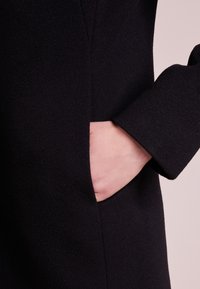 Detalje af sort uldfrakke med sidelomme, tekstureret stof og let udvidet ærmedesign. Simpelt og funktionelt design.