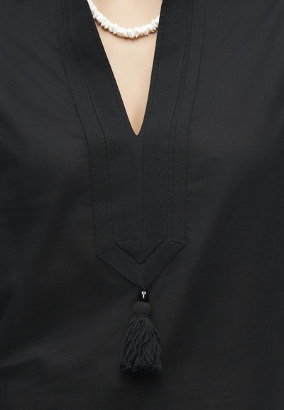 Μαύρο ένδυμα με V λαιμόκοψη με υφασμάτινη επένδυση και μαύρο φυλαχτό κρεμαστό κάτω από το λαιμό, φορεμένο με λευκό περιδέραιο από κοχύλια.