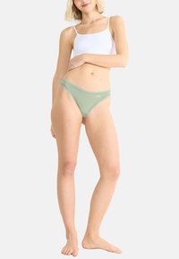 Lichtgroene vrouwen thong gemaakt van zachte stof, met een gladde textuur en delicate afwerking. Witte bralette erboven gedragen.