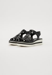 Des sandales compensées noires présentent des lanières en cuir entrecroisées, un design à bout ouvert et une semelle texturée blanche avec une bande noire en accent.