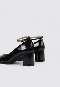 Escarpins en cuir verni noir avec un bout arrondi, une bride cheville et des talons bloc épais. Texture lisse avec une finition brillante.