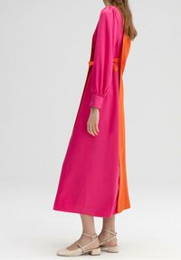 Femme debout de profil, portant une longue robe rose et orange à manches longues et des chaussures beiges à petit talon sur un fond uni.