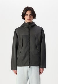 Svart hettejakke med to store frontlommer, glatt tekstur, glidelåsforing og elastiske ermer. Brukt med lyseblå jeans.