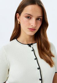 Geribbelde gebreide top met korte mouwen in crème met zwarte afwerking, voorzien van een ronde halslijn en een kleine knoopsluiting, accentueert met een strikdetail.