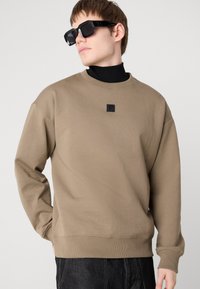 Rjava majica s kapuco, izdelana iz mešanice bombaža, z dolgimi rokavi, krožnim izrezom in majhno črno oznako na sredini prsnega dela. Model nosi črne sončne očala.