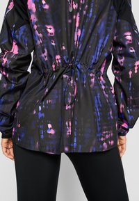 Veste noire avec un motif éclaboussé coloré en rose et bleu. Présente une taille cintrée avec un cordon de serrage ajustable et des poignets élastiques.