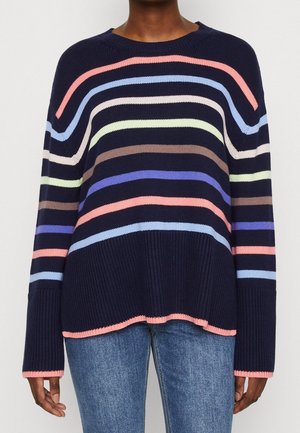 Personne portant un pull en tricot bleu marine avec des rayures pastel horizontales et un jean bleu clair, debout devant un fond uni.