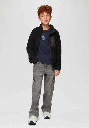 Schwarze Fleecejacke mit einer Reißverschlusstasche über einem marineblauen T-Shirt mit einem blauen Grafikdesign, kombiniert mit grauen Cargo-Hosen und weißen Sneakers.