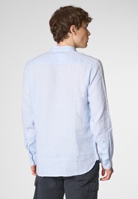 Un giovane con i capelli ricci che indossa una camicia a maniche lunghe blu chiaro è in piedi con le spalle rivolte verso l'osservatore, contro uno sfondo neutro.