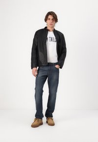 Zwarte leren jack met een rits aan de voorkant, witte grafische t-shirt, blauwe spijkerbroek en tan suède schoenen met een textuurzool.
