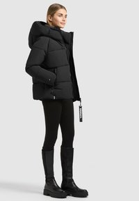 Femme portant une doudoune noire avec capuche, des leggings noirs et des bottes noires hautes jusqu'aux genoux, debout sur un fond clair uni.