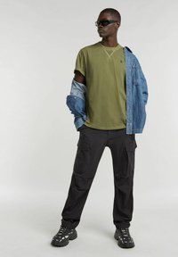 T-shirt verde oliva, giacca di jeans a vestibilità ampia, pantaloni cargo neri e calzature nere con accenti bianchi, che mostrano materiali resistenti e un design informale.