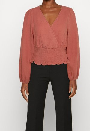 Vrouw draagt een roestkleurige blouse met lange mouwen, diepe V-hals en geplooide taille, gecombineerd met een zwarte broek.