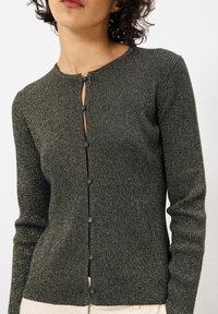 Dunkelgrüne, gerippte Strickjacke mit rundem Halsausschnitt, vorderer Knopfleiste und metallischen Fäden. Lange Ärmel und figurbetonte Passform.