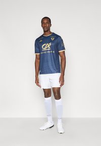 Tumsnējas zilas sporta formas krekls ar zelta akcentiem, ģeometrisku rakstu, īsām piedurknēm un "Centre France" zīmolu. Kopā ar baltām biksītēm un zeķēm.