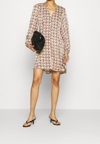 Femme en robe longue à manches fleuries tenant une pochette noire, portant des sandales à talons noires, se tenant devant un fond blanc uni.