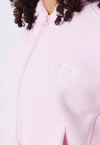 Haut à capuche zippé rose clair en tissu doux, comportant une étiquette de logo blanche et une poche avant pour le rangement.