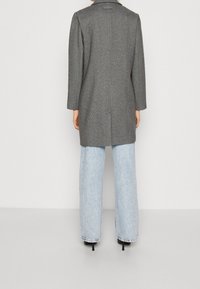 Abrigo gris de espiga con una silueta ajustada, dos aberturas traseras y mangas largas, combinado con unos vaqueros de pierna ancha azul claros y tacones negros.