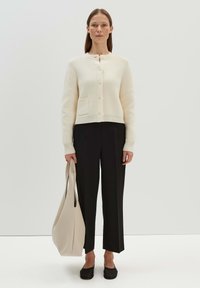 Cremefarbener Cardigan mit runder Ausschnitt, Knopfleiste und kleiner Tasche. Kombiniert mit schwarzen weiten Hosen und einer weichen beigen Tragetasche.