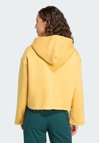 Felpa corta gialla con cappuccio grande, tessuto morbido e orlo non rifinito, abbinata a pantaloni verdi, che mostrano una vestibilità comoda e un design casual.