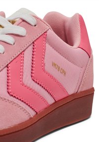 Tênis cor-de-rosa com um cabedal texturizado em tons variados de rosa, atacadores brancos e um logotipo dourado, montados sobre uma sola de borracha castanha.