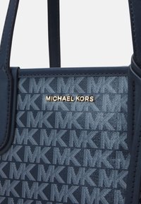 Gros plan sur un sac bleu Michael Kors avec un motif MK répétitif et le nom de la marque en or au centre, montrant des bretelles en cuir et des détails de couture.
