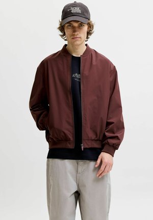 Jeune homme portant une veste bomber marron foncé, un t-shirt graphique noir, un pantalon gris et une casquette de baseball noire Jack & Jones, debout devant un fond blanc.