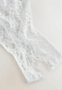 Tela de encaje blanco con elaborados patrones florales y bordes en forma de ondas, con una textura suave y translúcida y un corte asimétrico.