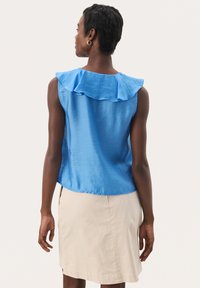 Haut sans manches en satin bleu avec un col à volants, au design fluide et à la texture lisse ; associé à une jupe beige longueur genoux.
