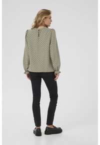 Olivgrön blus med svarta prickar, puffärmar, en nyckelhålsöppning i ryggen och volangärmar, kombinerad med svarta tajta byxor och skor.