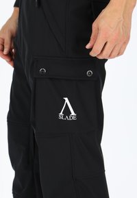 Zwarte cargobroek van flexibel materiaal met een zijzak met drukknoopsluiting en een prominent weergegeven wit logo "SLADE".
