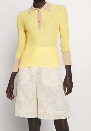 Person trägt einen gelben Strickpullover mit beigem Kragen und Bündchen, kombiniert mit cremefarbenen, strukturierten Shorts bis zum Knie.