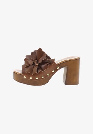 Brown leather heeled sandal with a large, layered floral embellishment and gold-tone studs along the base; chunky wooden heel.