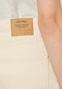 Jean en denim beige avec une étiquette en cuir affichant les instructions d'entretien et un logo. Taille de taille marquée comme 26, tissu texturé montré.