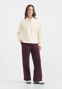 Kremowa koszulka polo z długim rękawem i guzikami, w połączeniu z szerokimi ciemnofioletowymi spodniami. Model stoi z rękami w kieszeniach, w szarych trampkach.