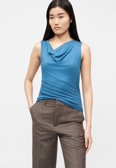 Νεαρή γυναίκα με μακριά σκούρα μαλλιά, ντυμένη με αμάνικο μπλε τοπ με ρίγες και καφέ tailored παντελόνι, στέκεται με το ένα χέρι στην τσέπη.