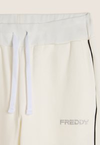 Pantalon de survêtement couleur crème avec une taille élastique blanche, des cordons de serrage blancs et un logo "FREDDY" argenté près de la hanche. Détail de bande latérale noire.