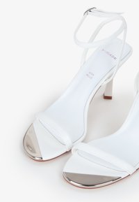 Hvide højhælede sandaler med et elegant design, der har tynde stropper og en metallisk tåkap. Lavet af læder med en glat tekstur.