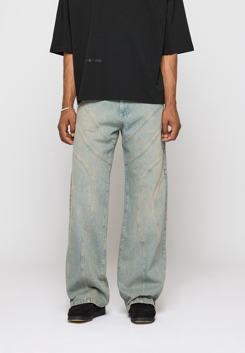 Personne portant un jean bleu clair à jambes larges avec des détails de coutures courbées, un t-shirt noir ample à manches courtes, des chaussures noires et un bracelet blanc.
