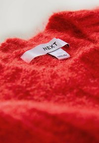 Červený huňatý svetr s jemnou texturou a viditelnou etikou s nápisem "NEXT." Látka vypadá nadýchaně a teple, zdůrazňuje svou bohatou barvu.