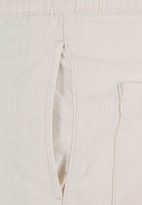 Gros plan sur un tissu beige montrant une poche fendue verticale avec des détails de couture et une partie d'une poche latérale rectangulaire.